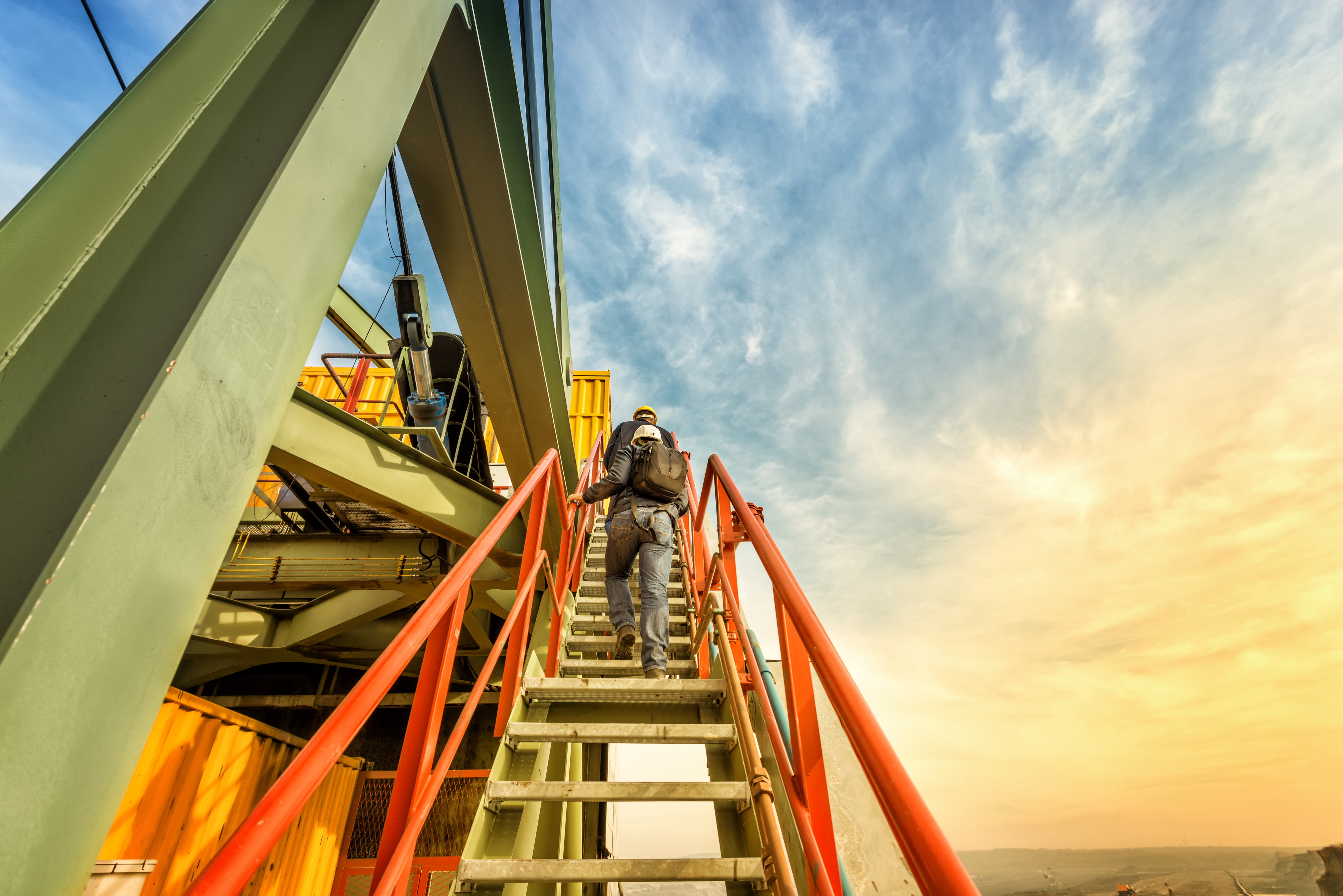 Two coal mine engineers are climbing on the steep stairs to the highest part of the huge coal digger machine. Beautiful and colorful sky in the background