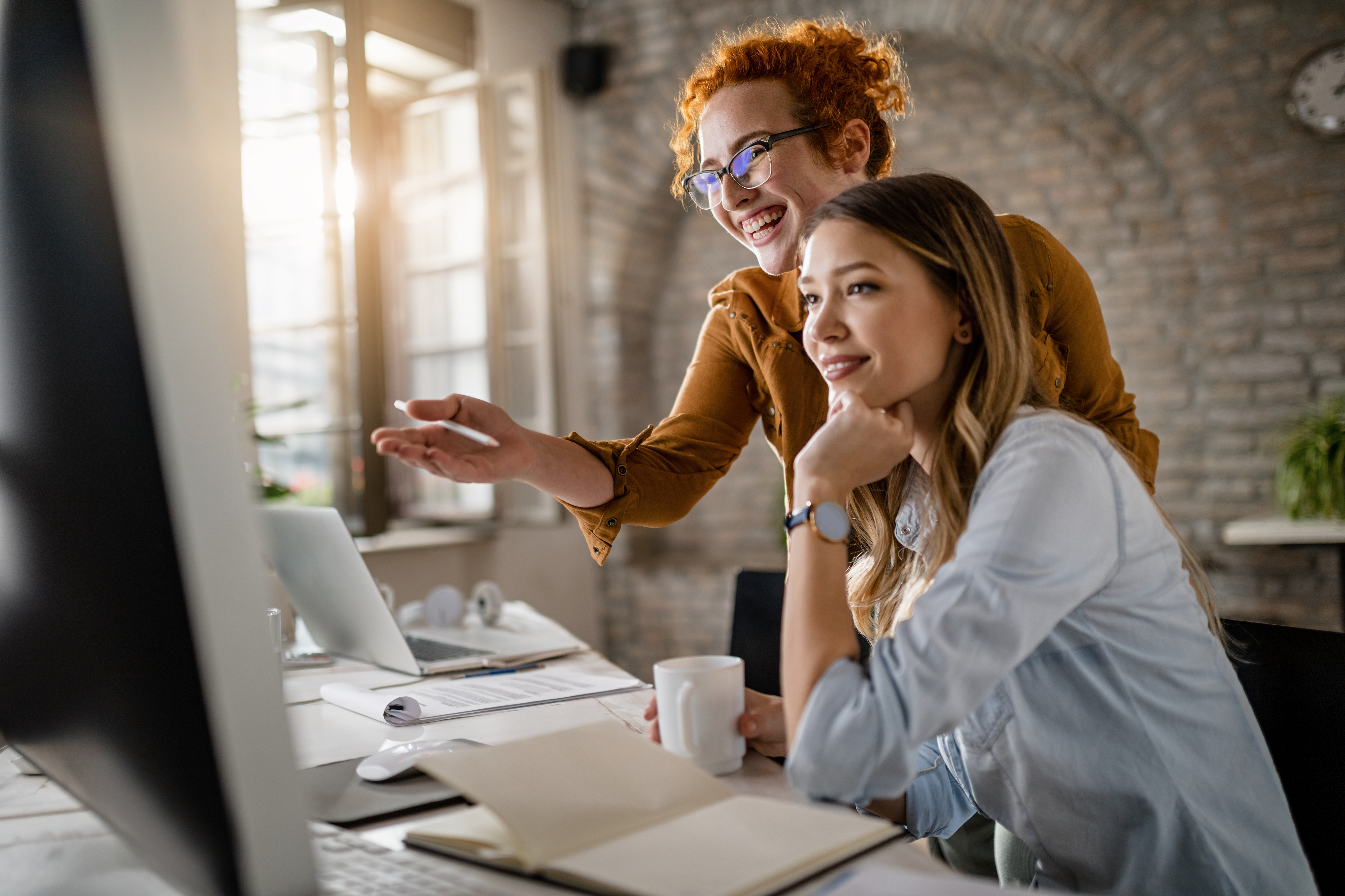 Young happy businesswomen cooperating while using desktop PC in the office